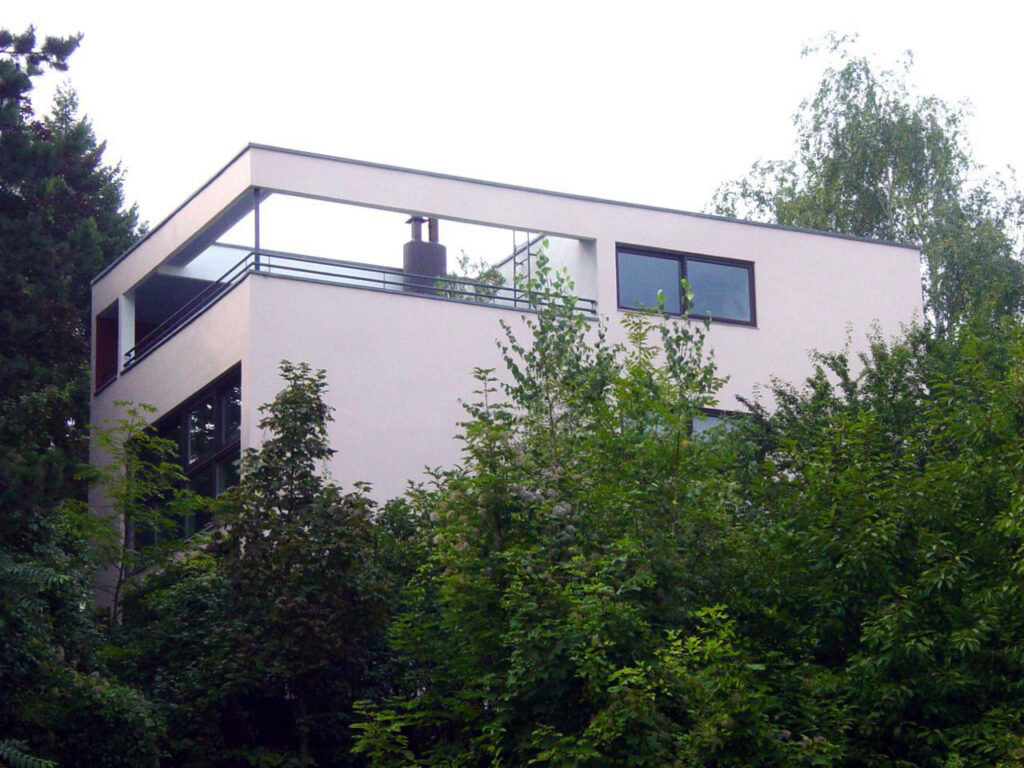 Modern minimalist house with large windows and balcony in a lush green setting.
