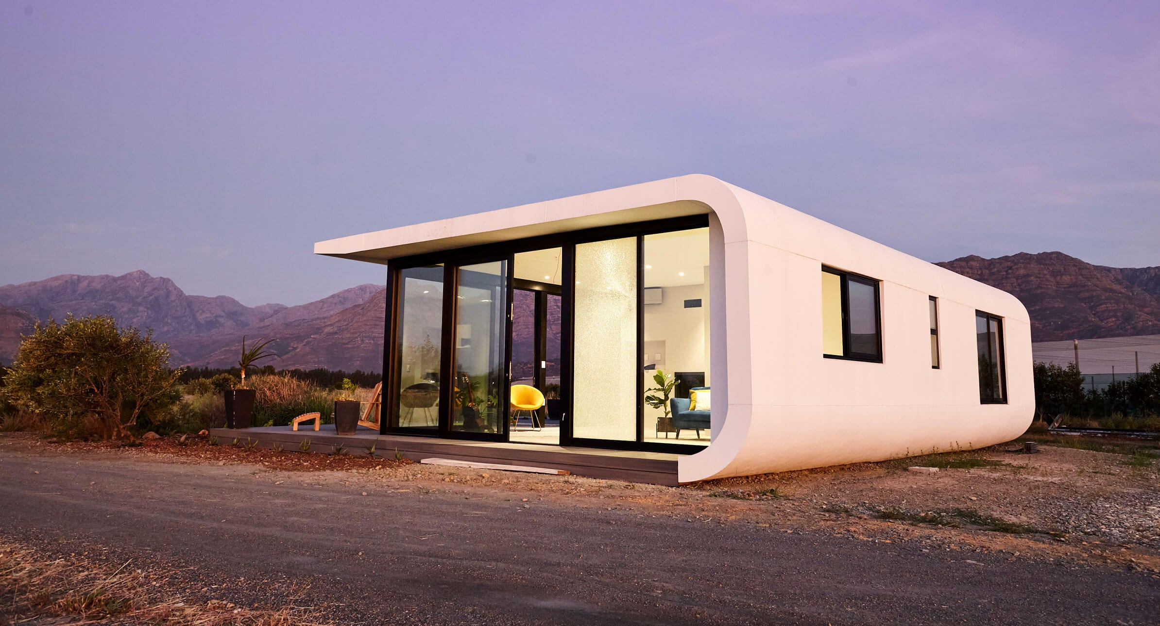 A modern, white 3D-printed house with large glass windows sits on a dirt lot, surrounded by greenery and mountains during sunset. The interior is warmly lit and minimalistic.