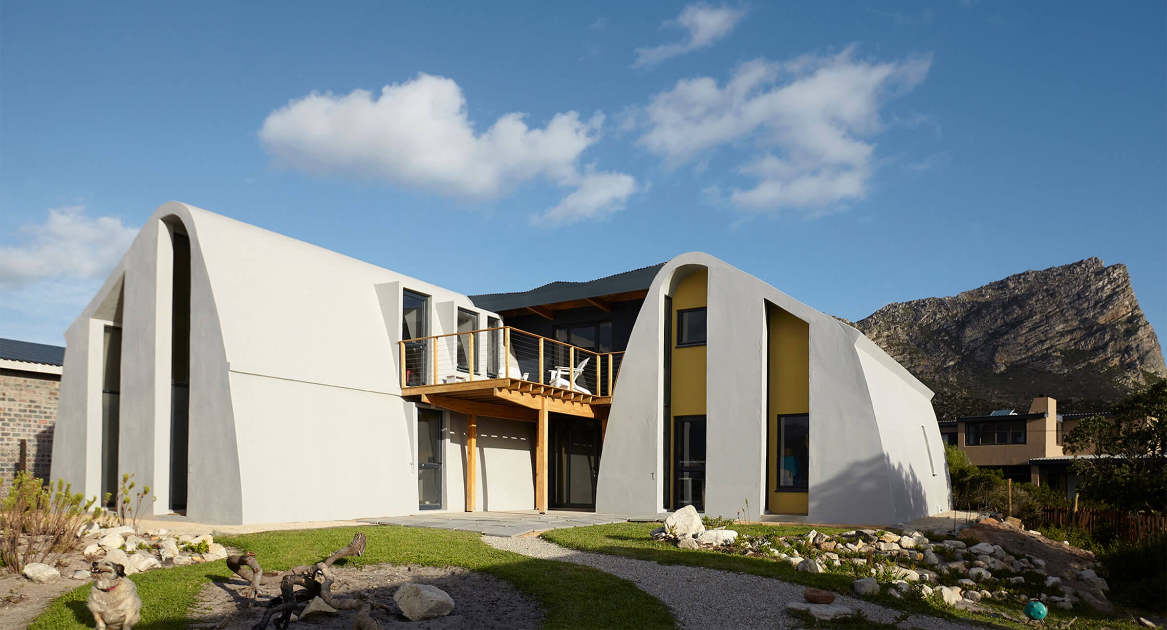 Modern two-story house with curved white walls, large windows, and a small balcony. The house sits on a grassy lawn with rocks and is set against a backdrop of mountains and a blue sky with scattered clouds.