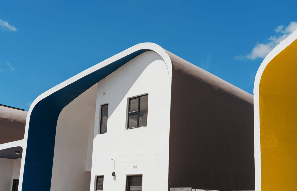 Modern white building with bold curved roof edges painted blue and yellow, set against a clear blue sky with minimal clouds. Large windows and clean lines give it a contemporary, minimalistic look.