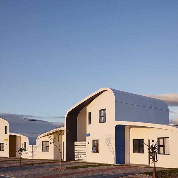 A modern white house with a curved roof and blue accents, featuring large windows and minimal exterior decor. The scene is lit by soft sunlight under a clear sky.
