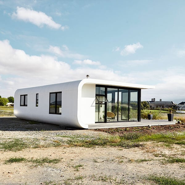 Modern white, rectangular house with rounded corners, large glass windows, and a flat roof, sitting on a bare, grassy plot under a partly cloudy sky.