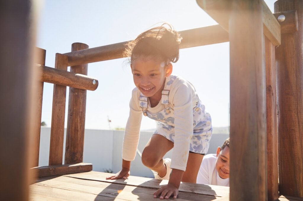 Climbing on wooden playground structure with smiling girl, outdoor play area, sunny day.