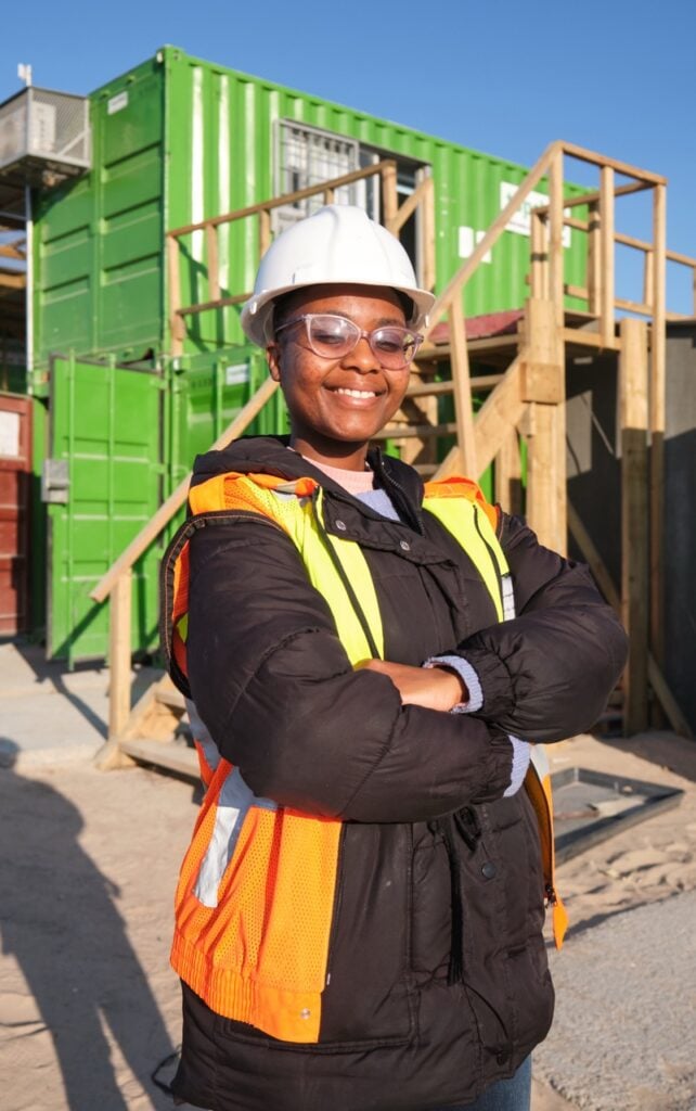 Female construction worker in safety gear at a building site with green shipping containers.