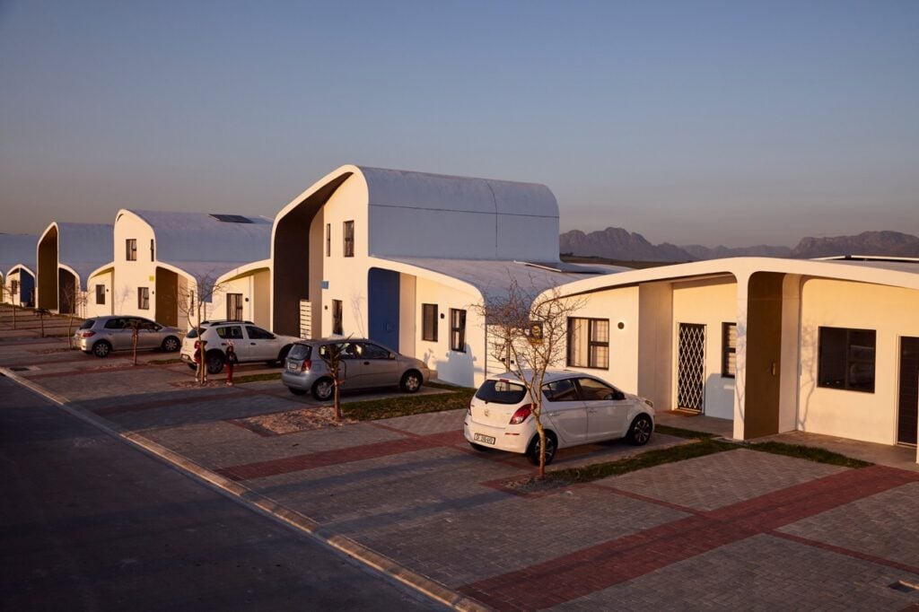 An exterior photograph of a row of modern white Citra eHomes captured at golden hour, featuring distinctive sweeping curved roofs, paved driveways with parked cars, and mountains in the background under a soft evening sky.