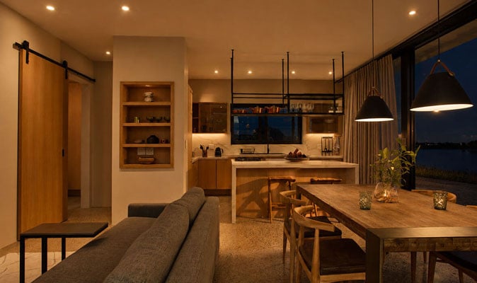 Warmly lit modern kitchen and dining area with wooden furniture, open shelves, pendant lights, and large windows overlooking a dark evening view outside. Cozy and inviting atmosphere.