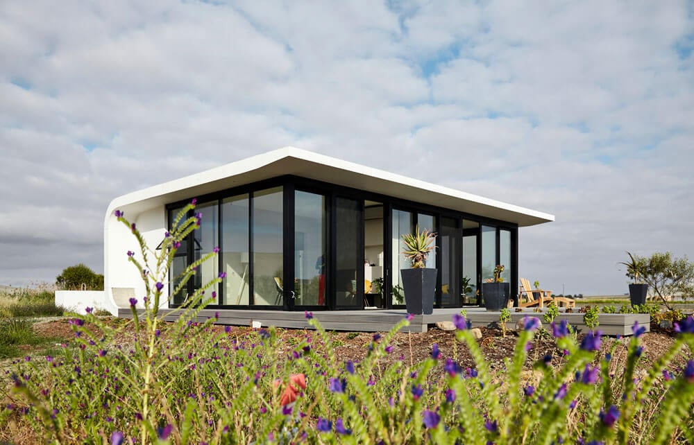 Contemporary glass house featuring large windows and a sleek design, set amidst lush garden landscap.
