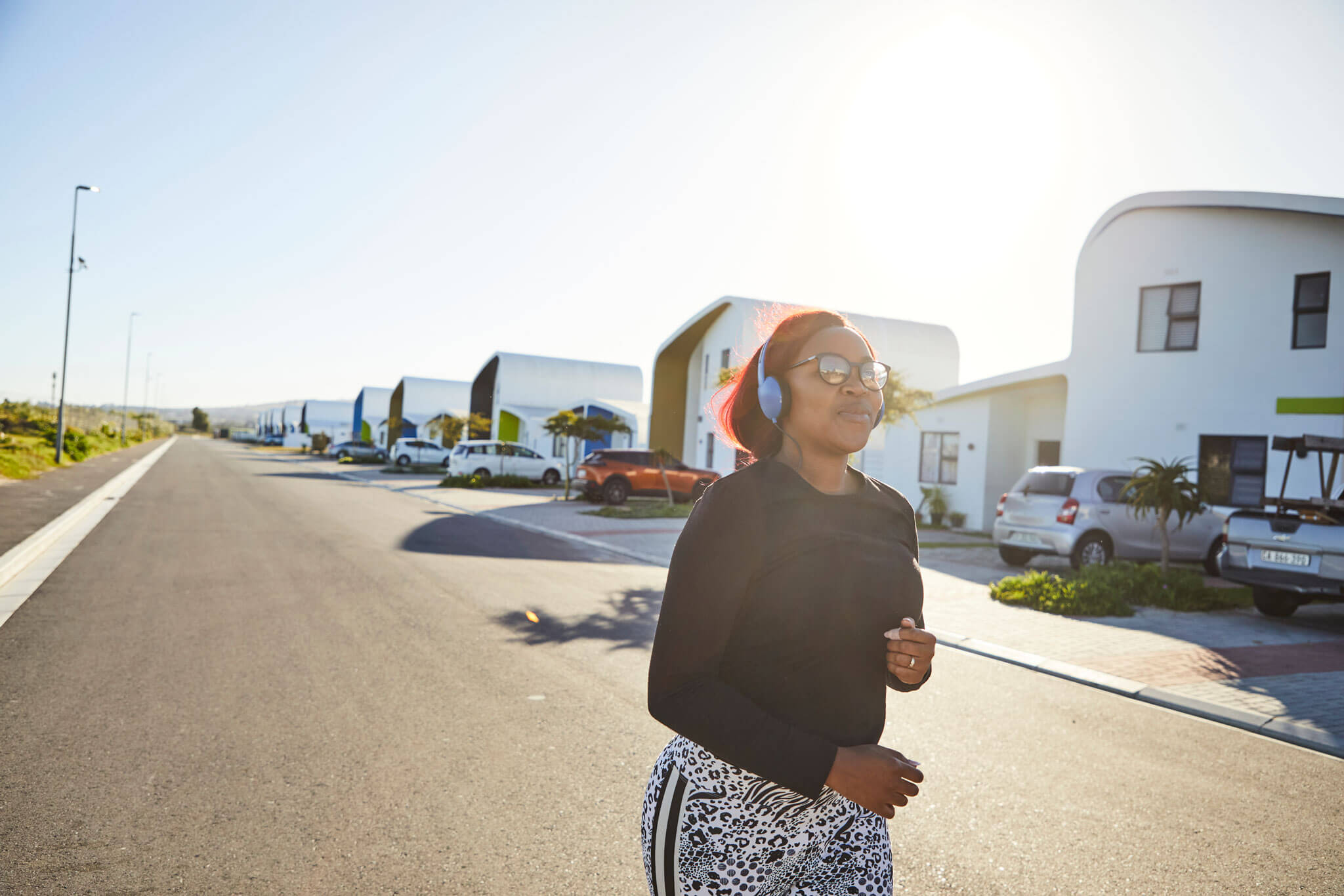Neighbourhoods A woman with headphones and sunglasses jogs down a sunny, quiet suburban street lined with modern houses and parked cars.