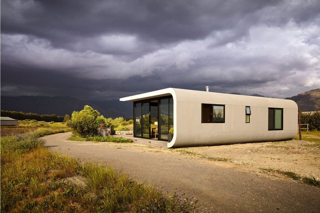Modern minimalist tiny house with curved design in a rural landscape under stormy sky.