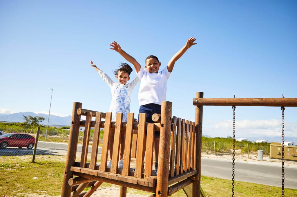 Two smiling children stand on a wooden playground platform with arms raised high against a bright blue sky.