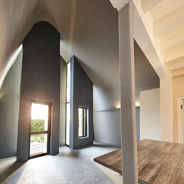 Modern, minimalist interior with high vaulted ceilings, large tall windows, smooth grey walls, and a wooden table; sunlight streams through the open door, creating a bright, airy atmosphere.
