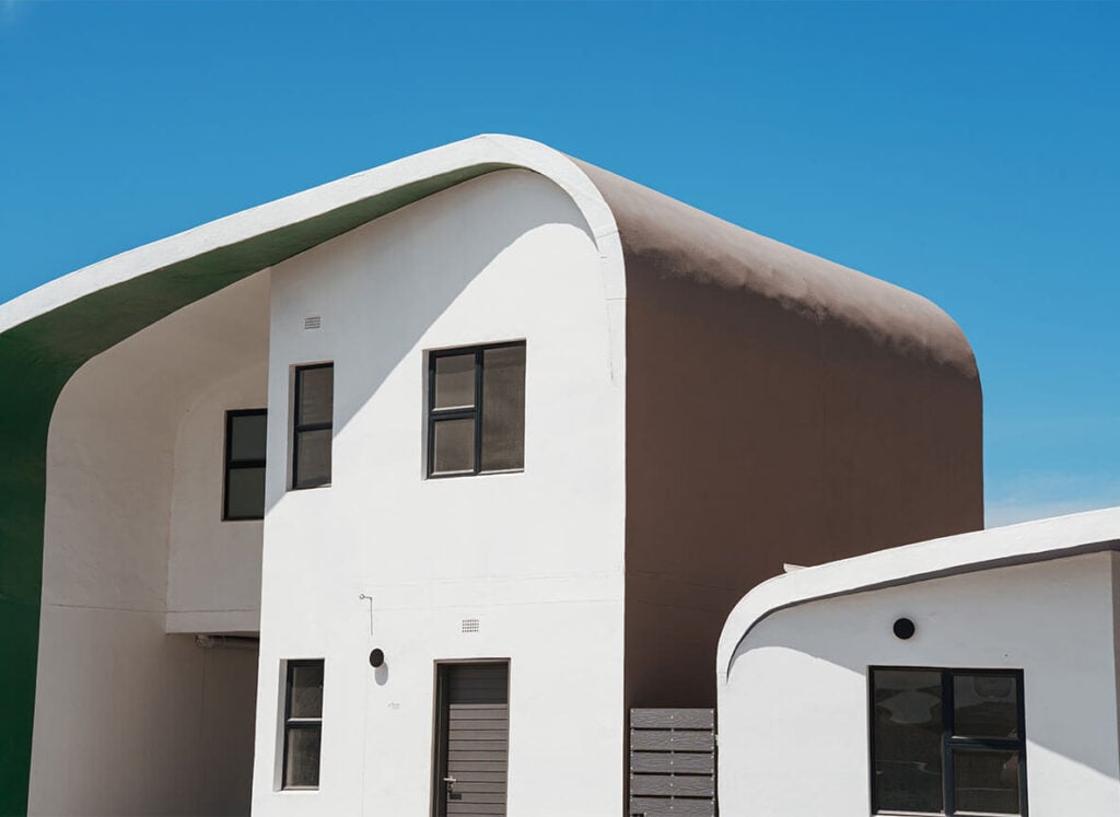 Modern white building with a curved brown roof and green overhang, next to a smaller white building, under a blue sky.