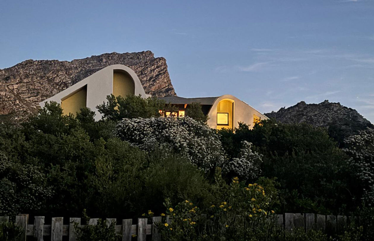 Sustainability Modern curved building with illuminated windows nestled in green bushes, with a rocky mountain behind under a twilight sky.