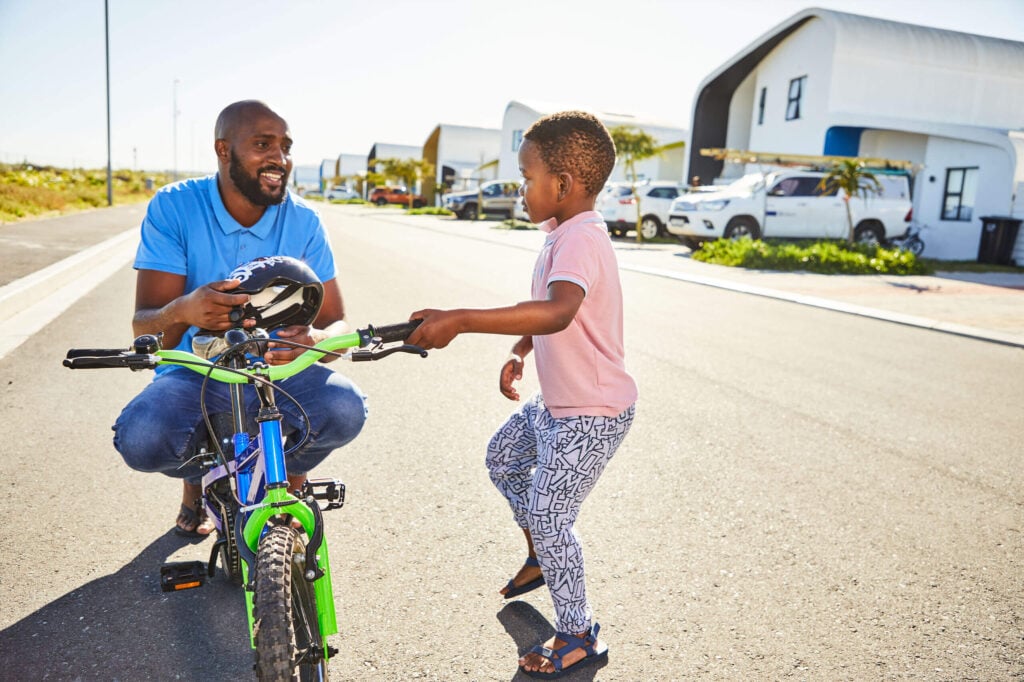 Man crouching, holding a helmet, smiles at a child holding a bicycle's handlebars on a sunny street.