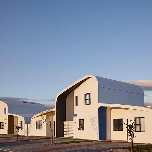 A modern white house with a curved roof and blue accents, featuring large windows and minimal exterior decor. The scene is lit by soft sunlight under a clear sky.