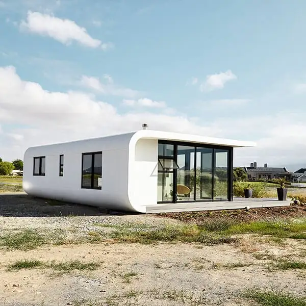 Modern white, rectangular house with rounded corners, large glass windows, and a flat roof, sitting on a bare, grassy plot under a partly cloudy sky.