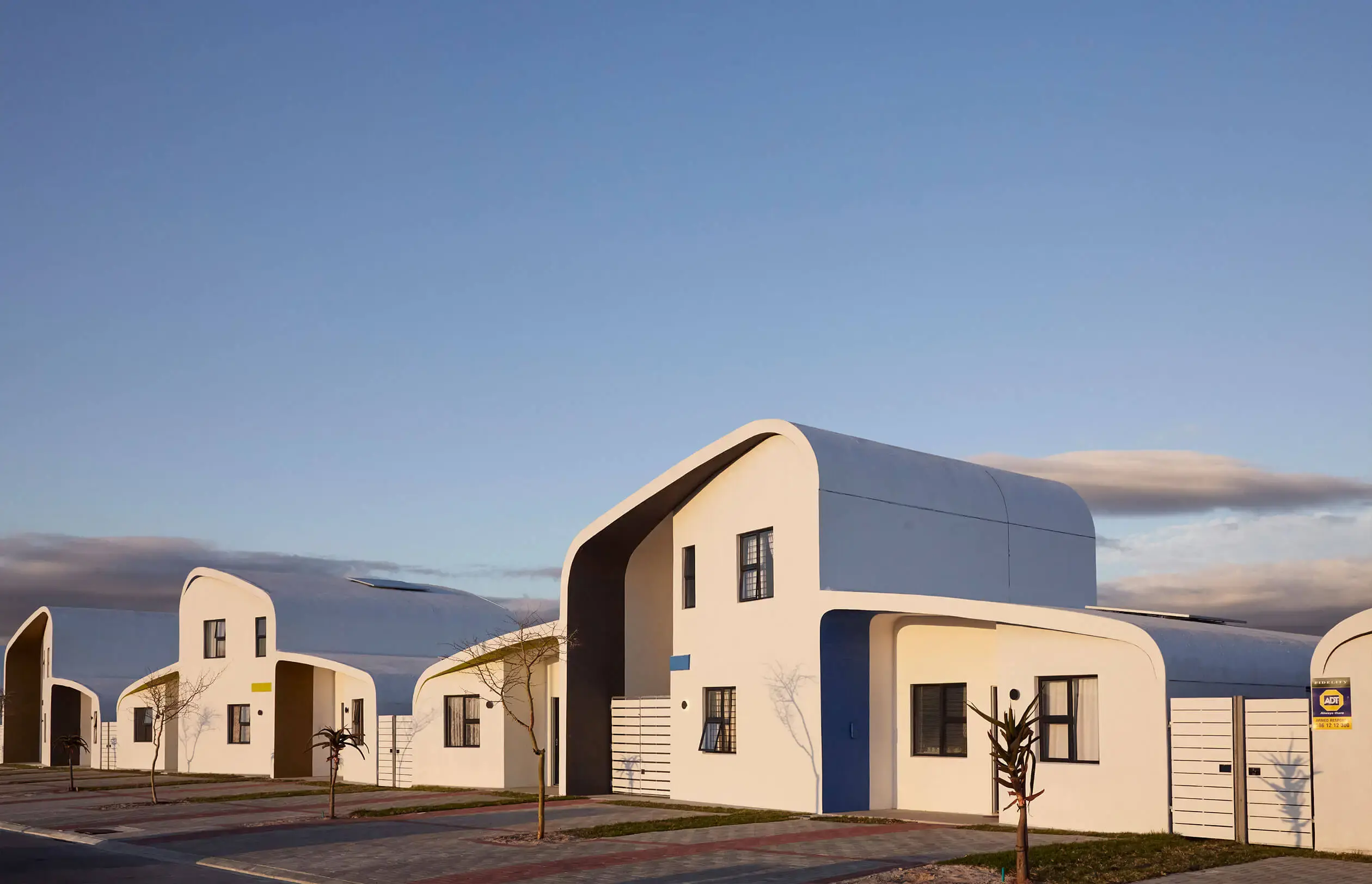 Modern white houses with curved roofs and large windows line a quiet street under a clear sky at sunset. Sparse young trees and a paved sidewalk are visible in front of the homes.