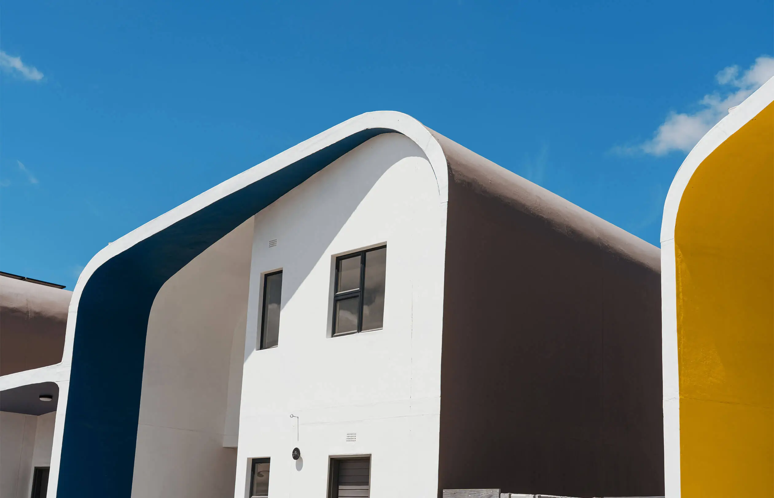 Modern white building with bold curved roof edges painted blue and yellow, set against a clear blue sky with minimal clouds. Large windows and clean lines give it a contemporary, minimalistic look.