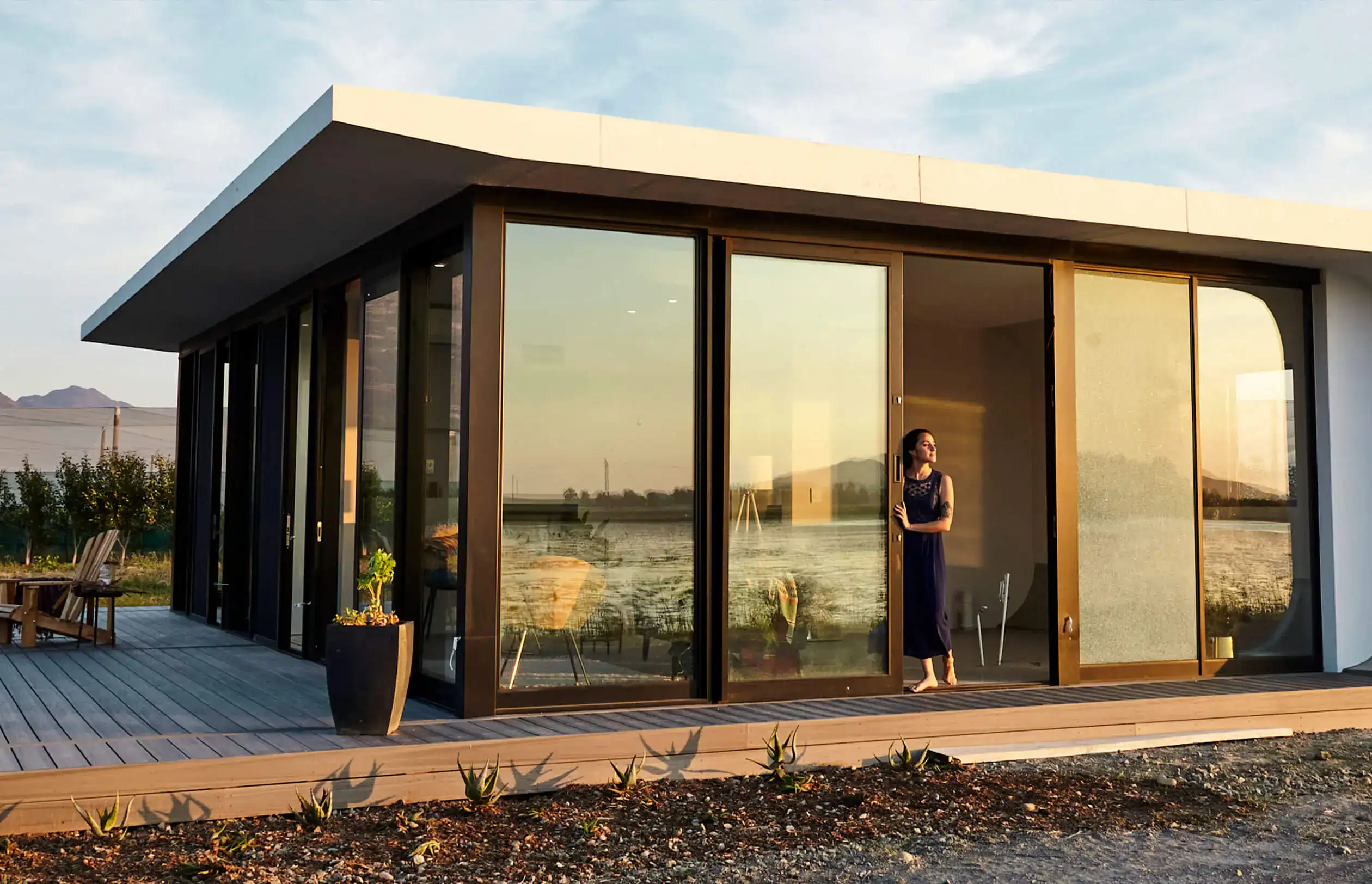 A woman stands near the sliding glass doors of a modern house with large windows, wooden deck, and outdoor chairs, overlooking a body of water and mountains at sunset.