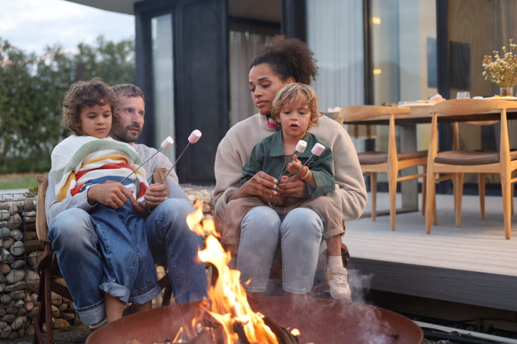 A family of four sitting around an outdoor fire pit at dusk, with two adults holding young children on their laps as they roast marshmallows over the flames in front of a modern Citra home.
