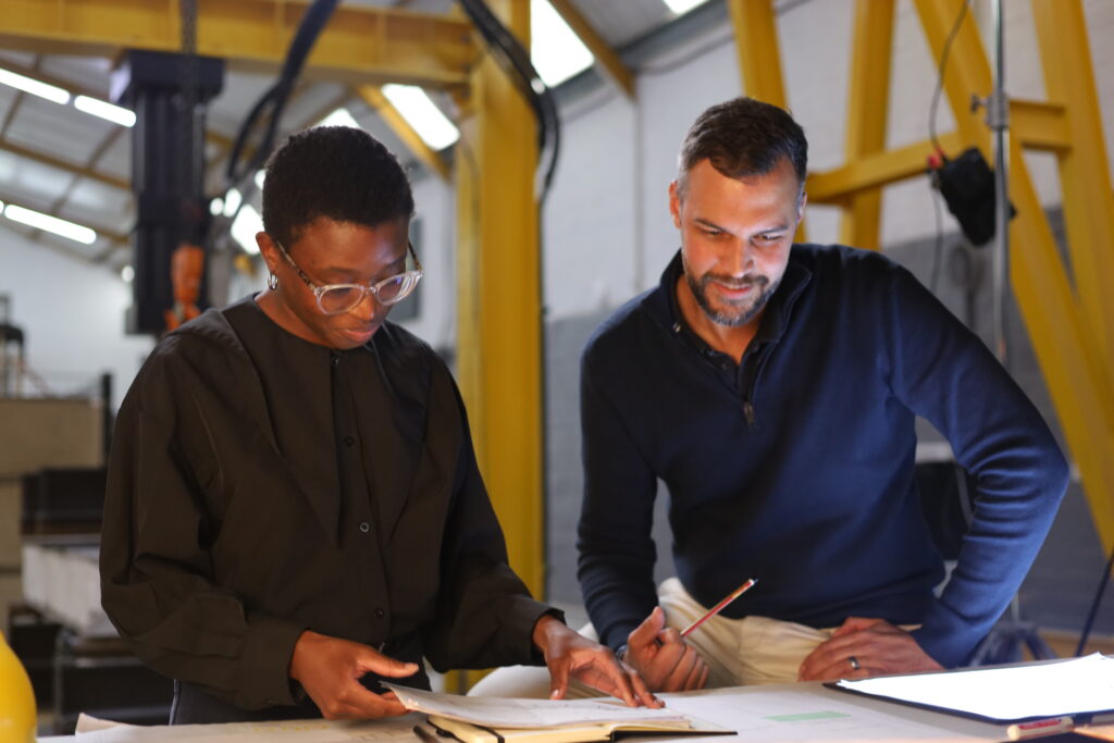 An indoor shot of two Citra team members collaborating over building plans laid out on a wide workstation. One colleague points to a section of the blueprint with a pencil while holding reference papers, as the other leans in closely to inspect the drawings. In the background, factory machinery and yellow crane structures are visible under bright ceiling lights.