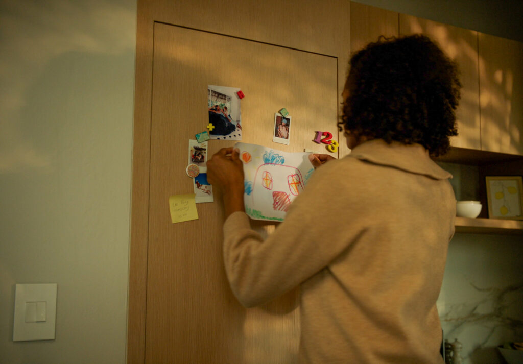 A mother pinning a colorful child's drawing of a house onto a wooden refrigerator panel alongside family photos, a sticky note, and colorful magnets in a warm kitchen setting.