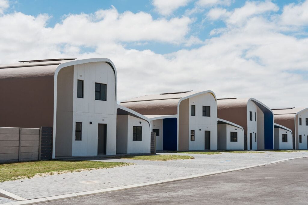 A street-level view of newly built smart sustainable homes with distinctive curved brown shell roofs topped with solar panels. The modern two-story housing units feature clean white facades, dark-framed windows, and paved walkways under a bright sky with light clouds.